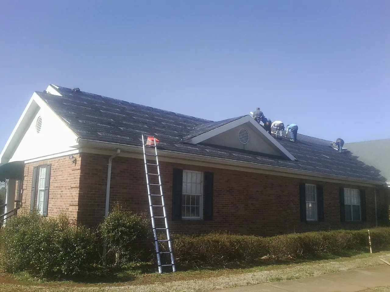 Crew tearing shingles off a brick ranch roof with a ladder set — Metal Roof Repair work by our crew in Richardsville