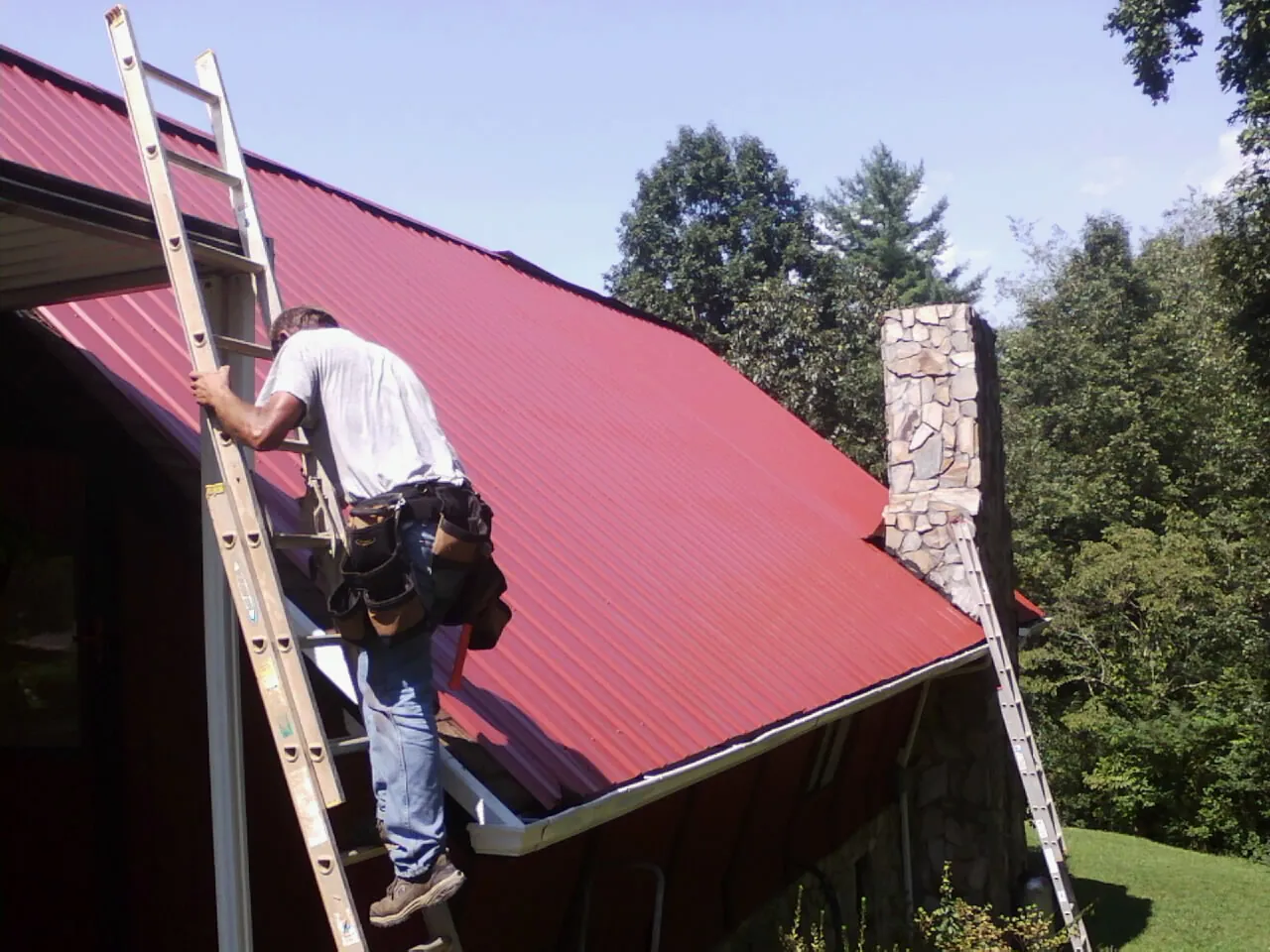 Roofer on a ladder beside a red metal roof and stone chimney — Soffit & Fascia Repair by Roofing Brothers in Richardsville